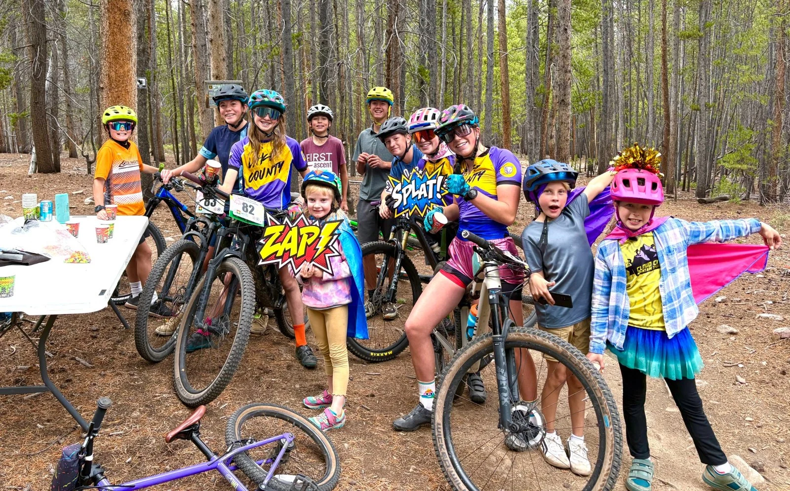 Adults and children in colorful costumes gather with their bikes in a wooded area.