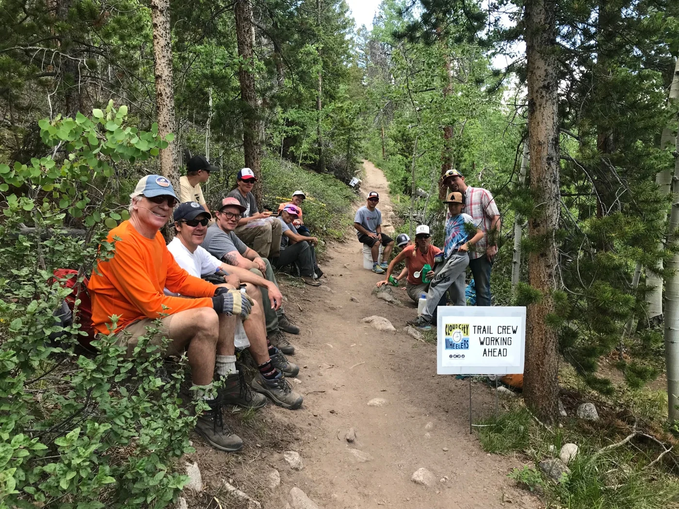 A volunteer trail crew takes a break beside a Cloud City Wheelers trail-work sign in the forest.