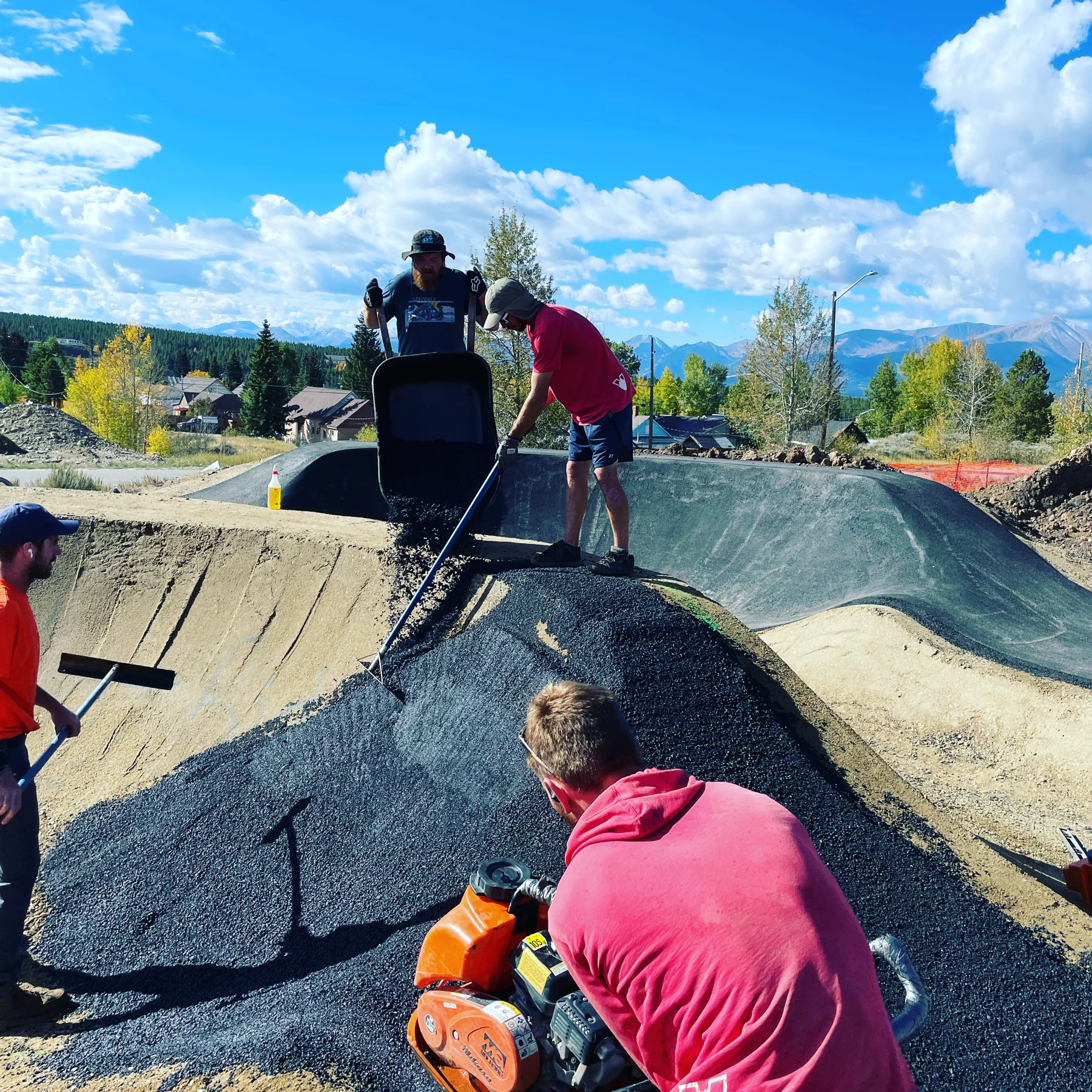 Crew members rake and compact an asphalt berm at Huck Finn Bike Park with mountains behind.