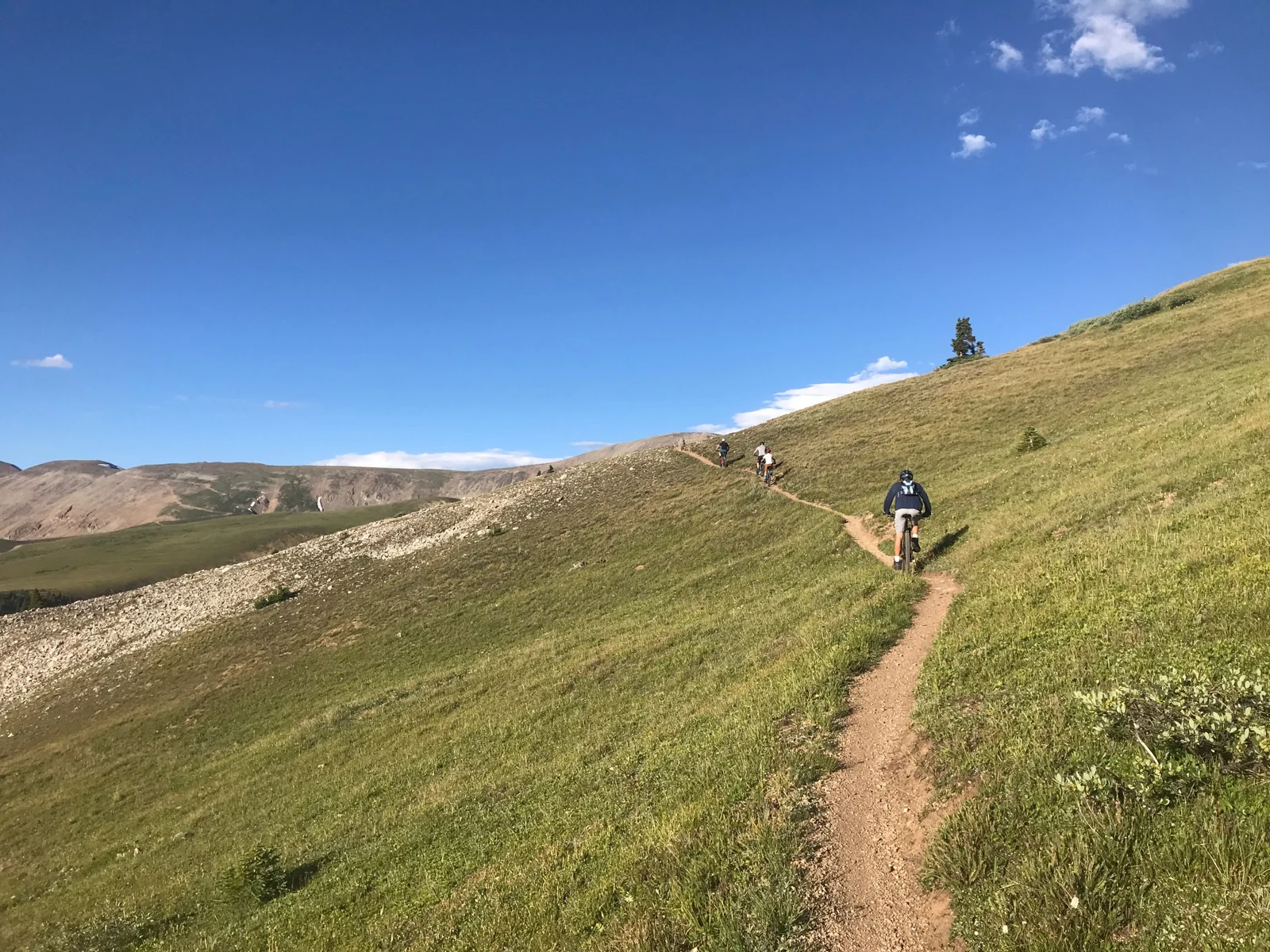 Cyclists follow a narrow singletrack across an open alpine hillside beneath a bright blue sky.