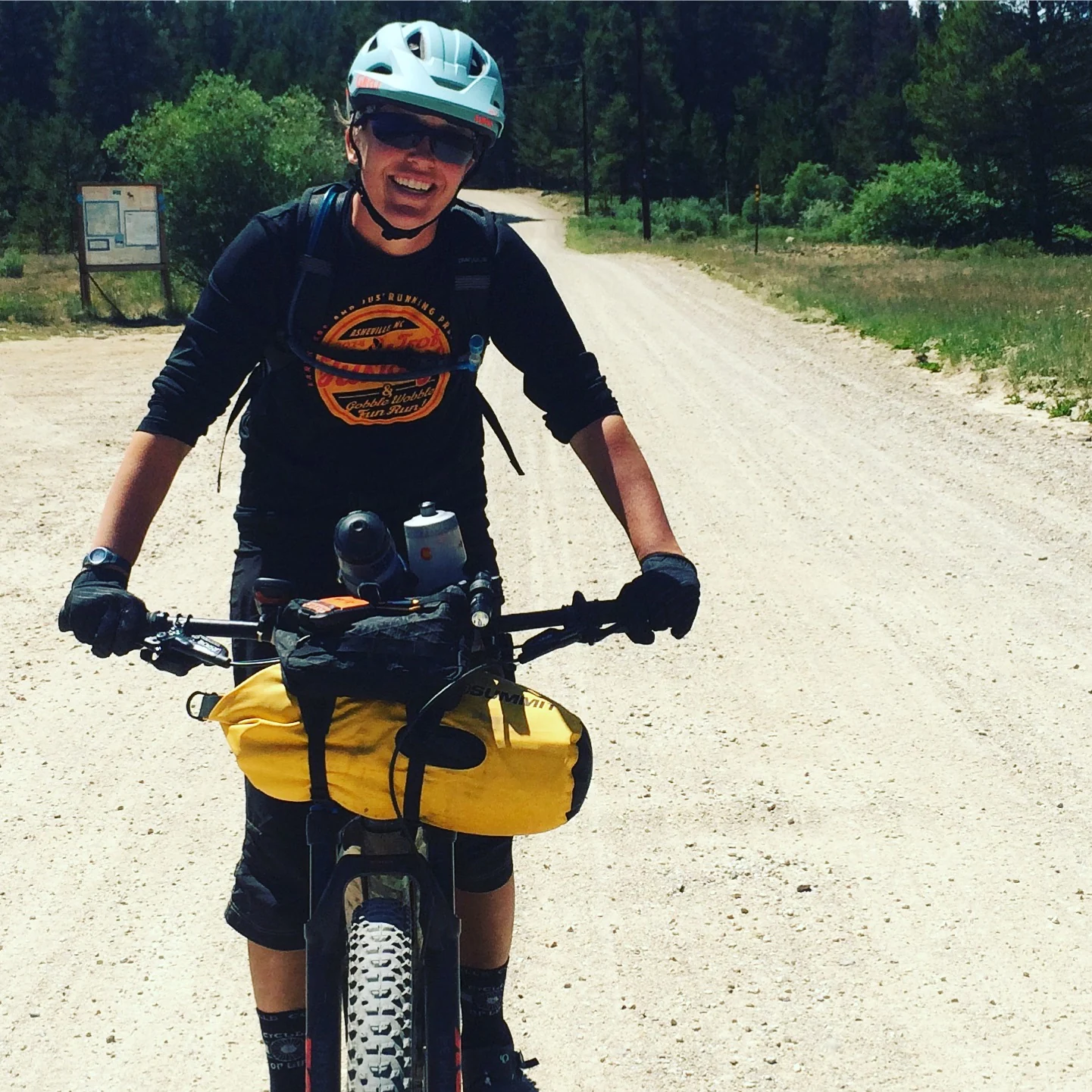 A smiling cyclist in a pale blue helmet pauses on a gravel road with a yellow bag on the handlebars.