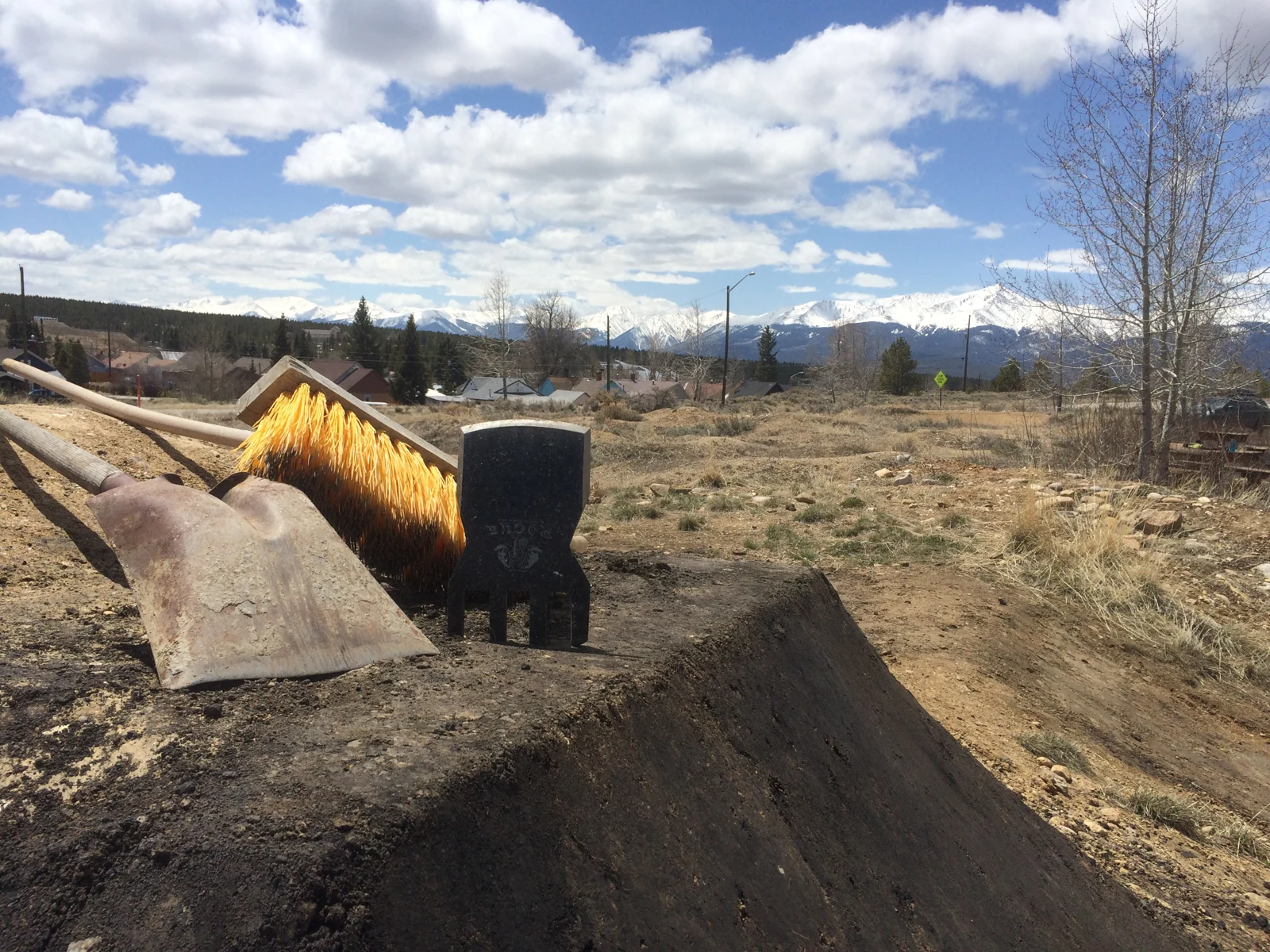 A shovel, yellow broom, and trail-building tool rest on a dirt berm with snowy mountains in the distance.
