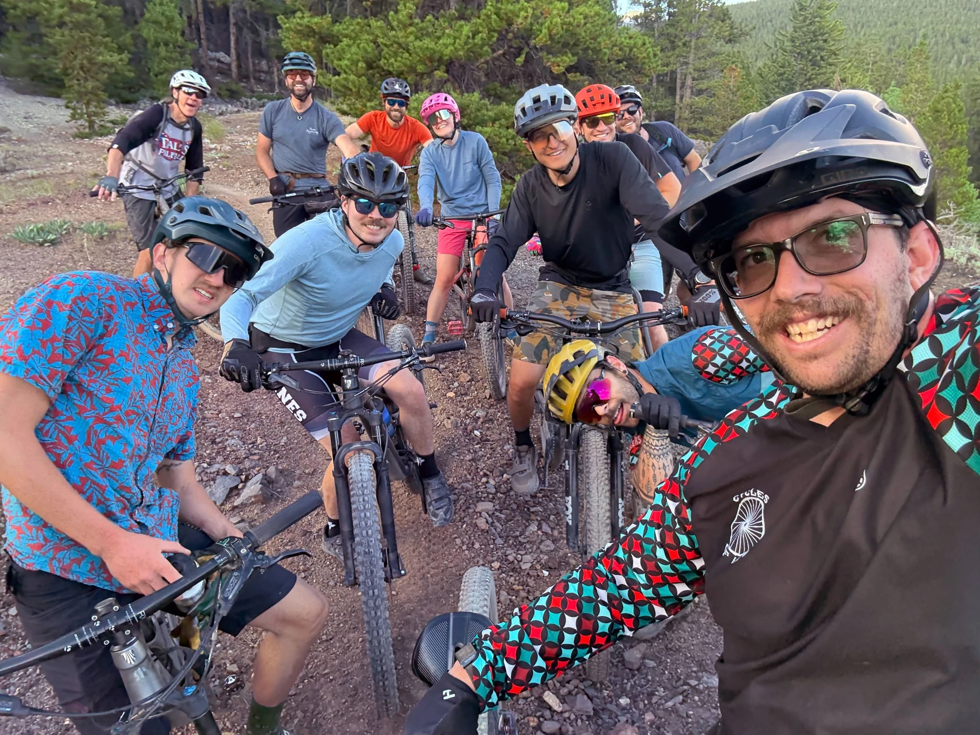 A group of smiling cyclists gathers with their bikes on a wooded trail.