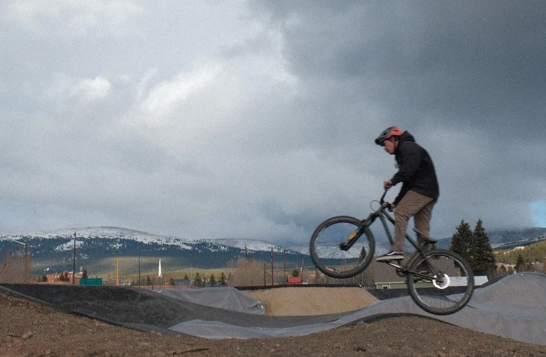 Cyclist jumping over asphalt pump-track mounds with mountains behind.