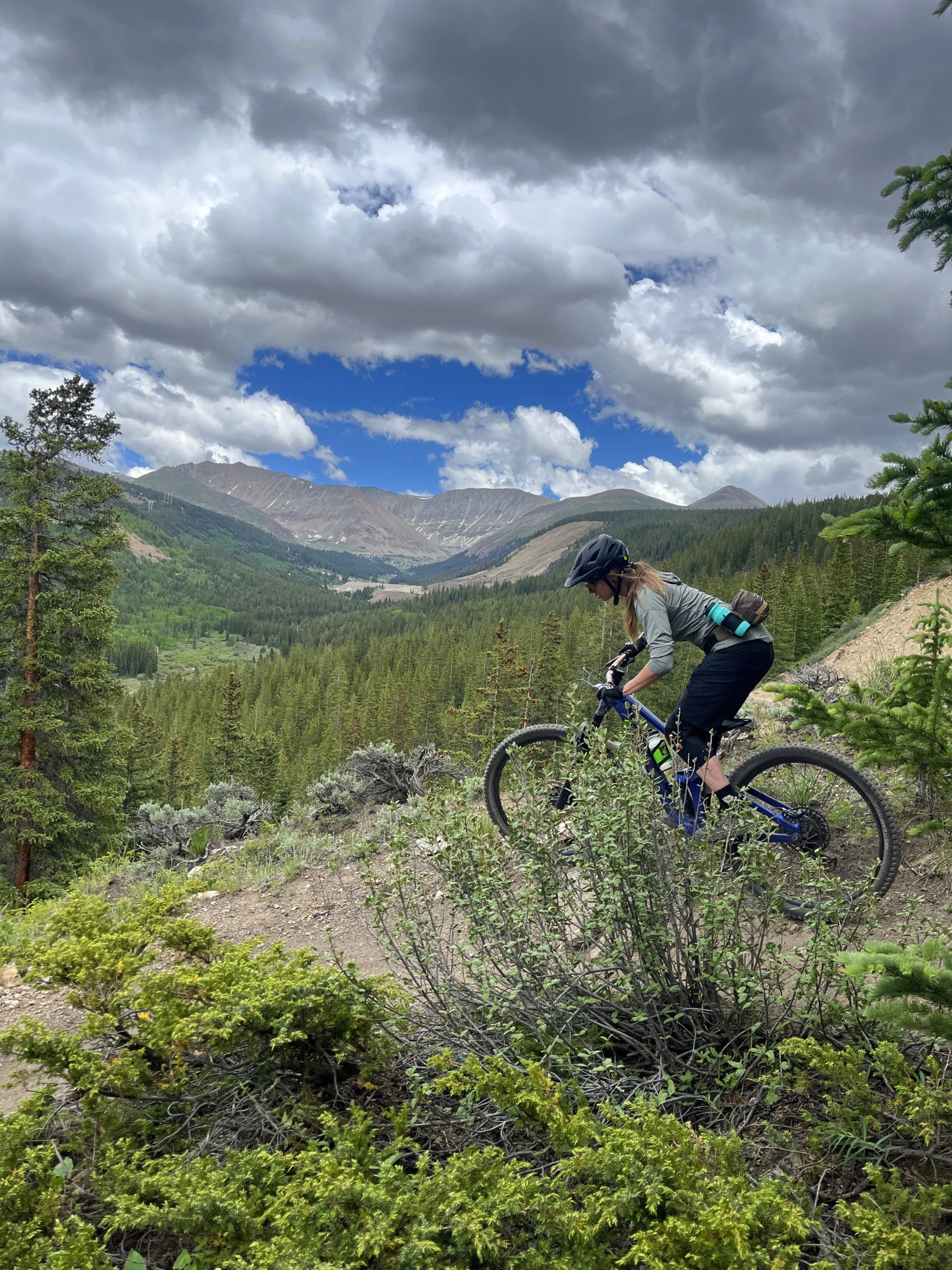Jenna riding a mountain trail above a green valley, with alpine peaks and dramatic clouds in the background.