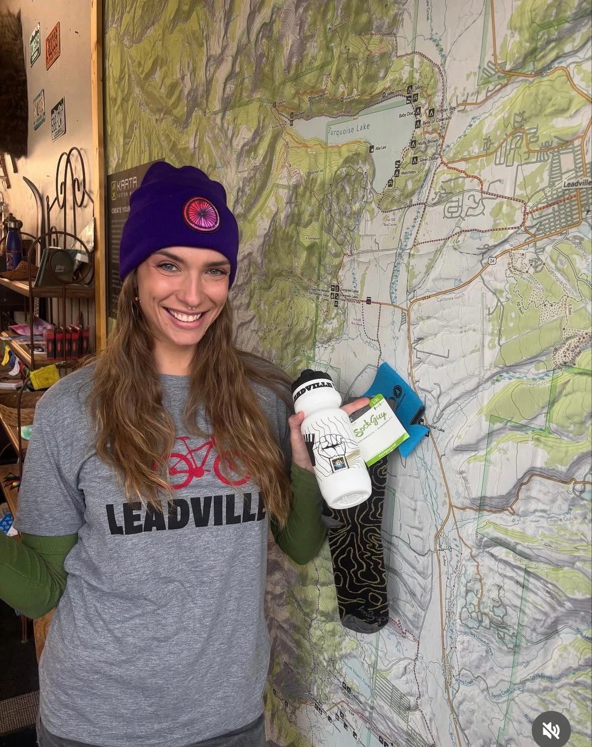 Jenna smiling with her photo-contest prizes, wearing a purple Wheelers beanie and Leadville shirt in front of a trail map.