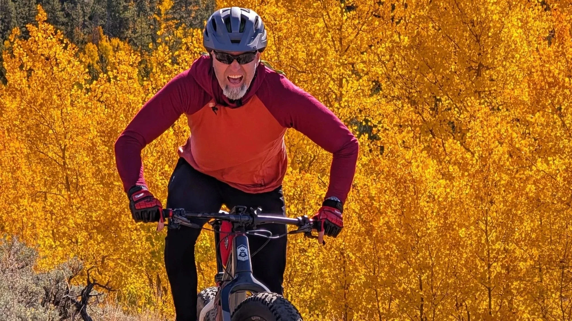 A smiling mountain biker in an orange and burgundy jersey rides in front of golden aspens.