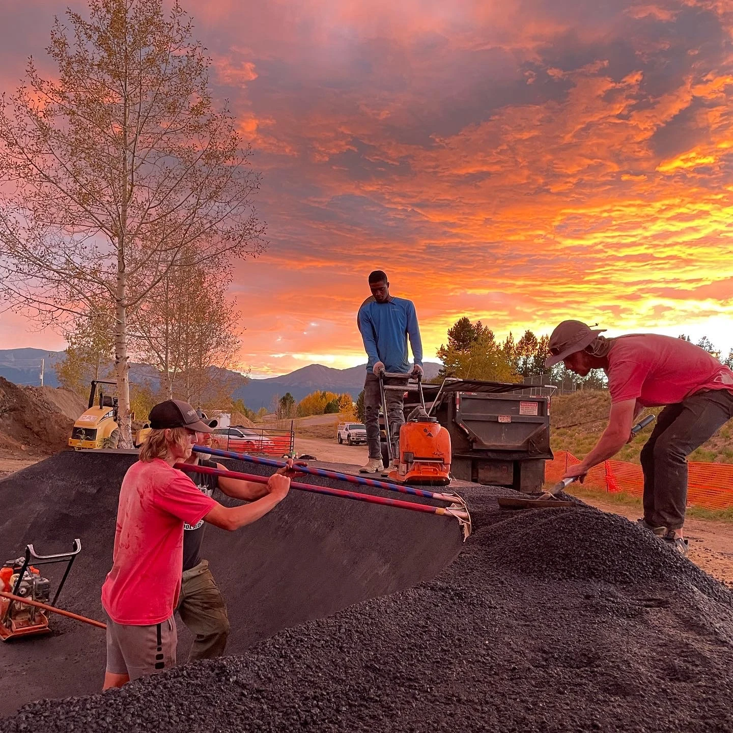 Crew raking and compacting pump-track asphalt under vivid orange sunset.