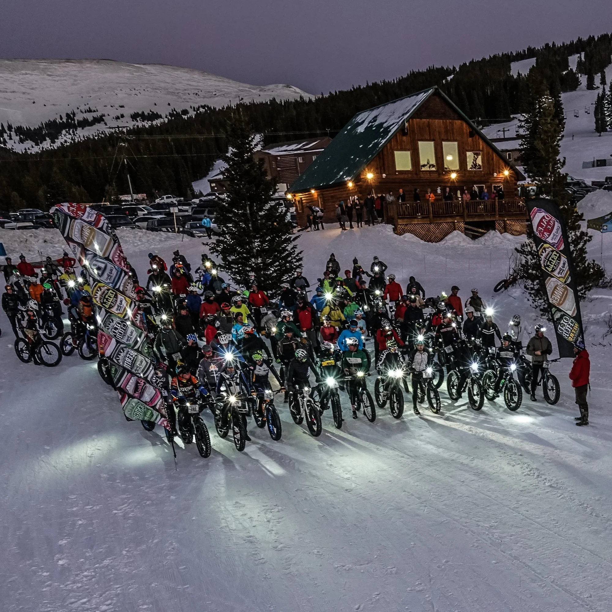A large Tennessee Pass Night Jam field gathers at dusk outside the lodge, with bike lights shining on the snow.