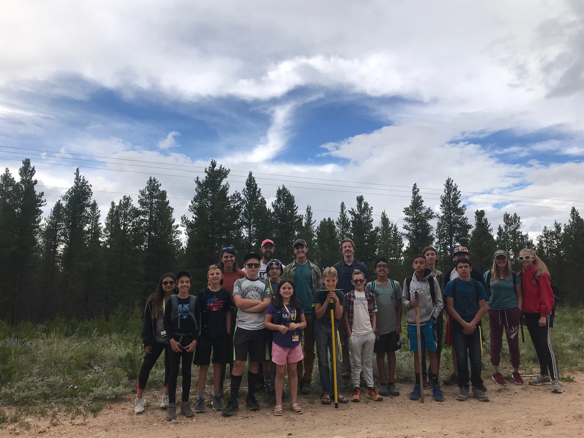 Large trail crew holding tools on trail beneath mountain clouds.