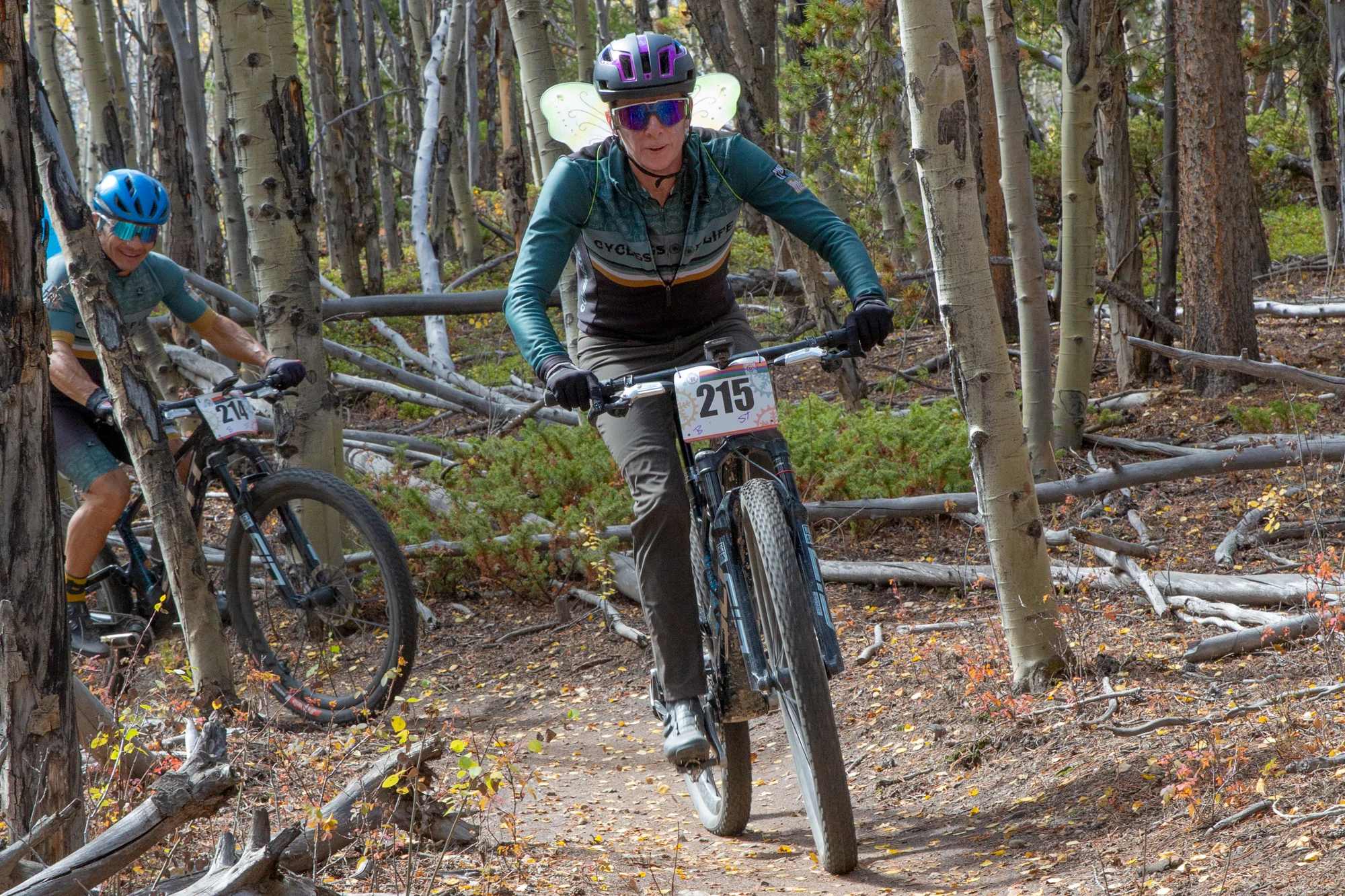 Two cyclists wearing fairy wings ride toward the camera on aspen-lined singletrack.