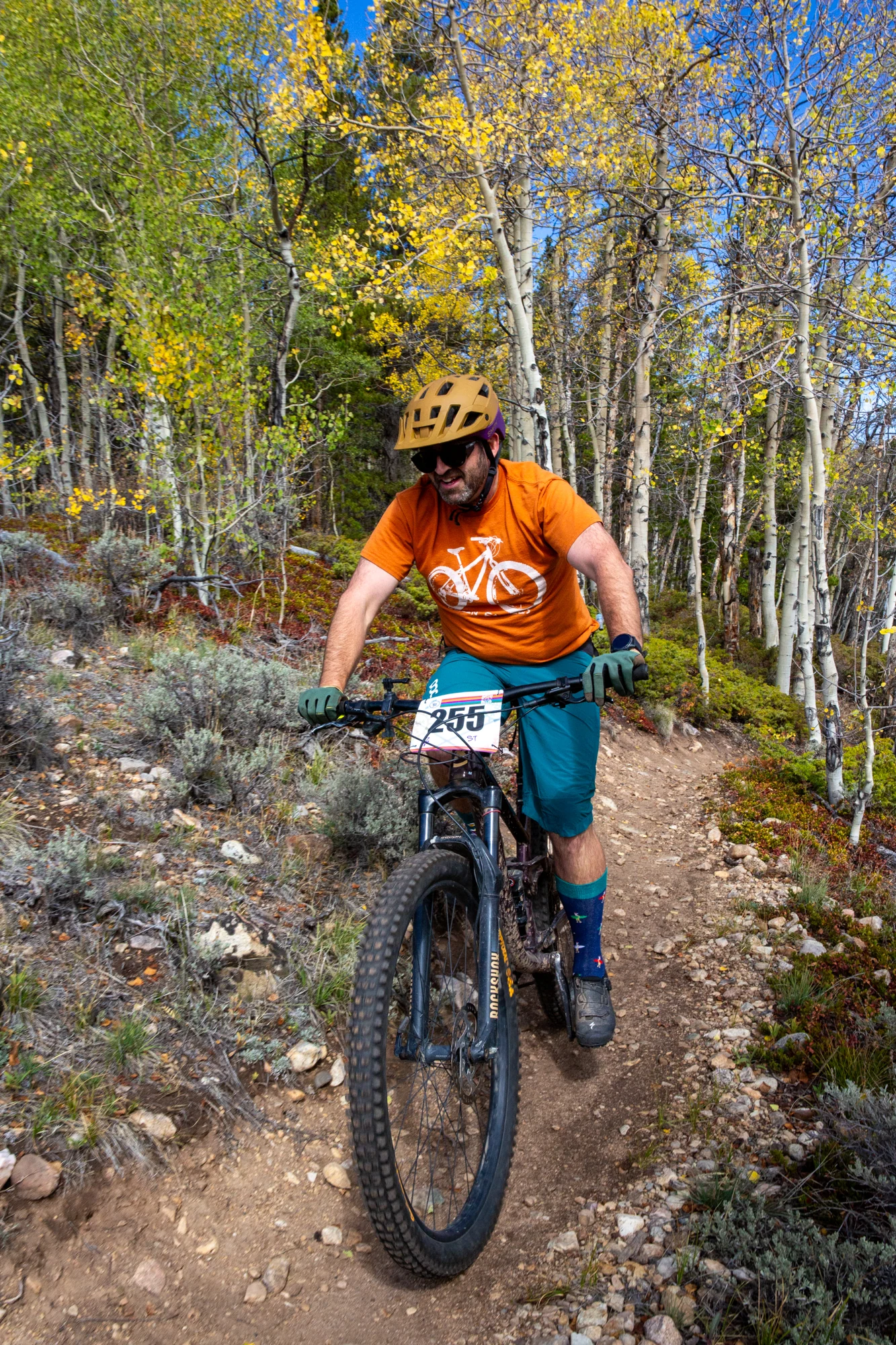 A smiling cyclist in an orange shirt rides toward the camera through golden aspens.