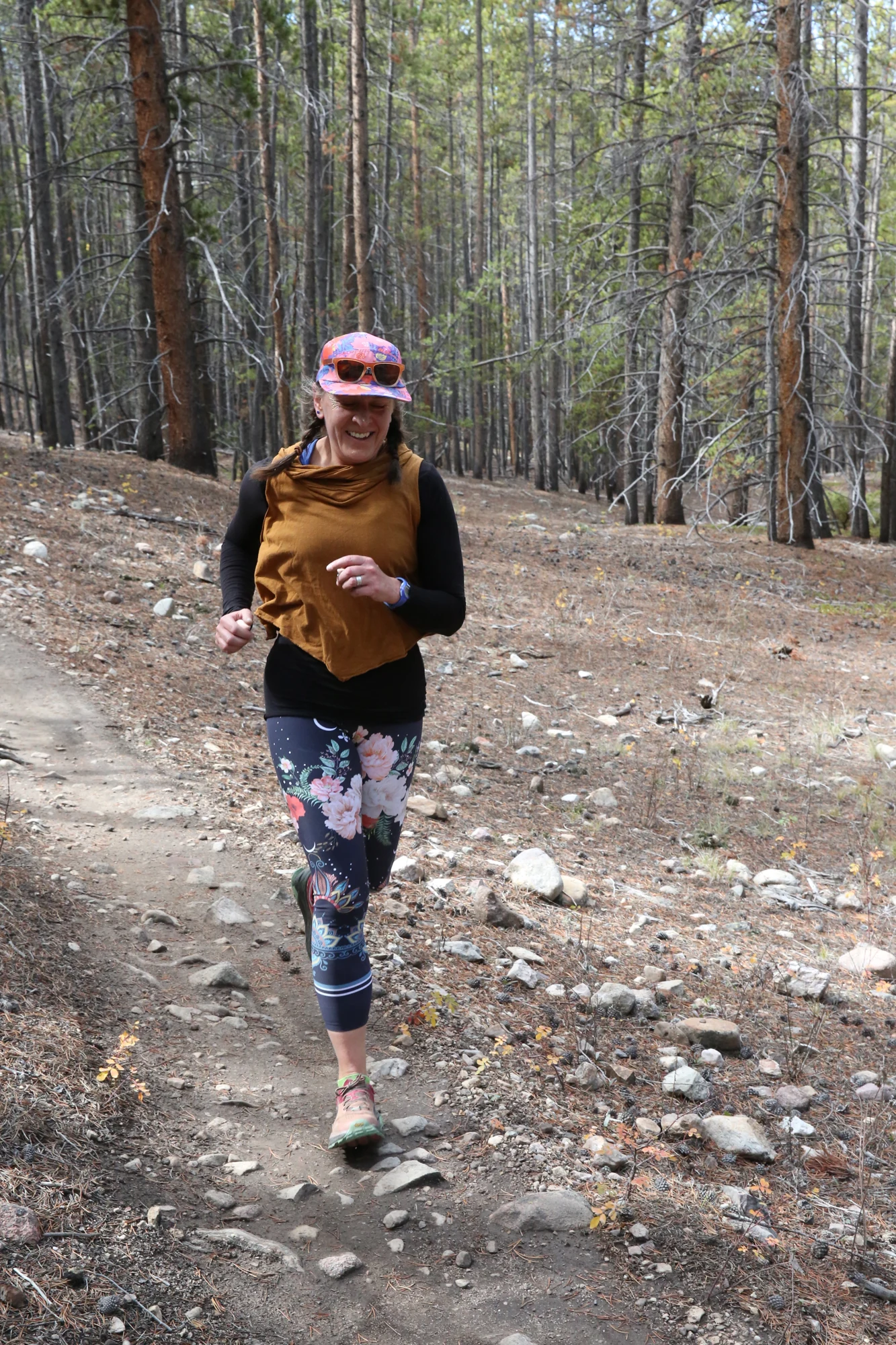 A participant runs toward the camera on a forest trail, wearing floral leggings and a pink cap.